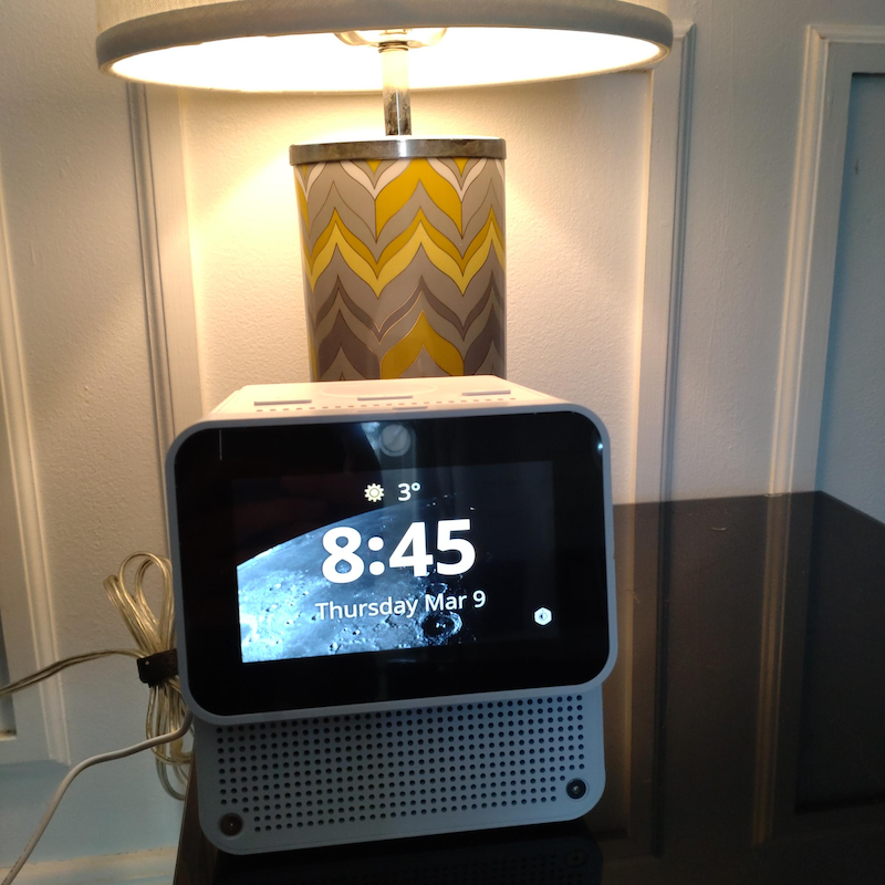 A white box with screen showing the time and a picture of the moon, and a table lamp behind it.